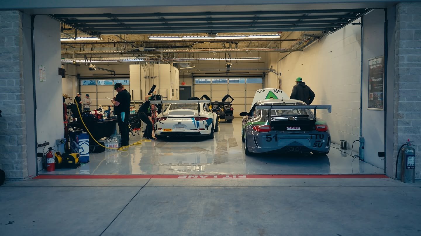 Two Porsche race cars in the pit lane garage with the team working