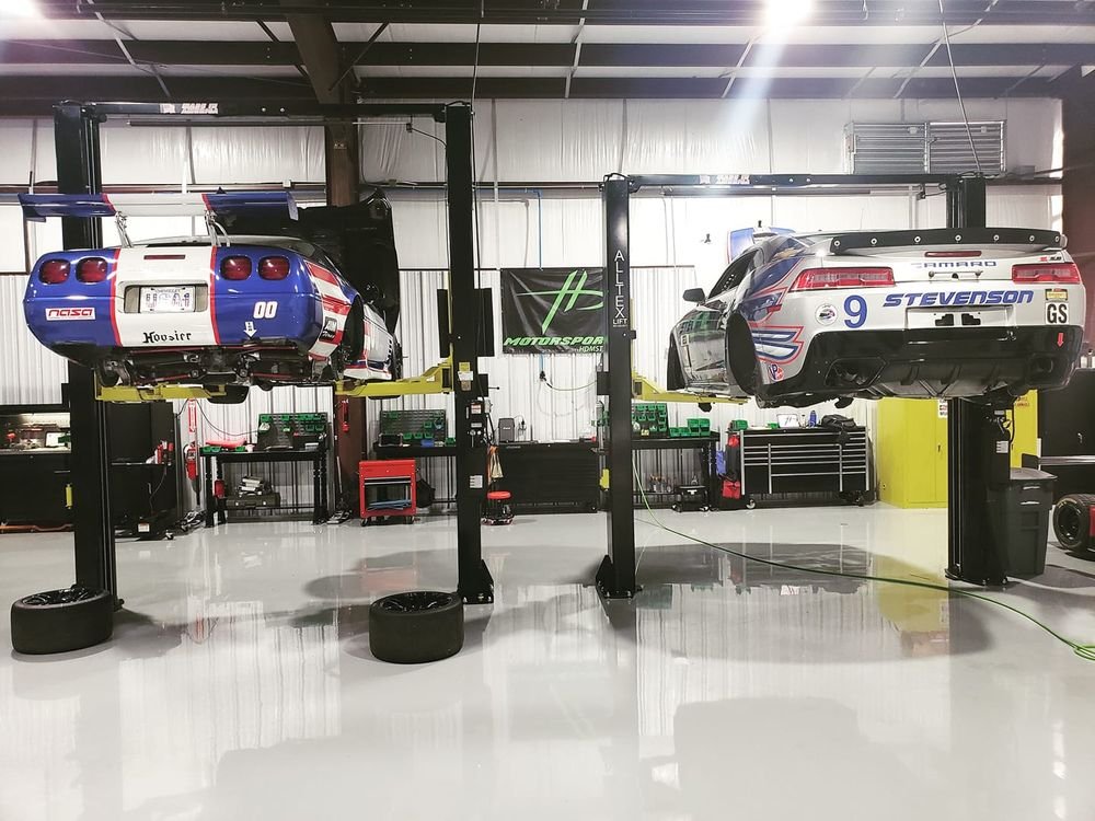 Two race cars on lifts in the HD Motorsports bay, a NASA Corvette and a Stevenson Motorsports Camaro